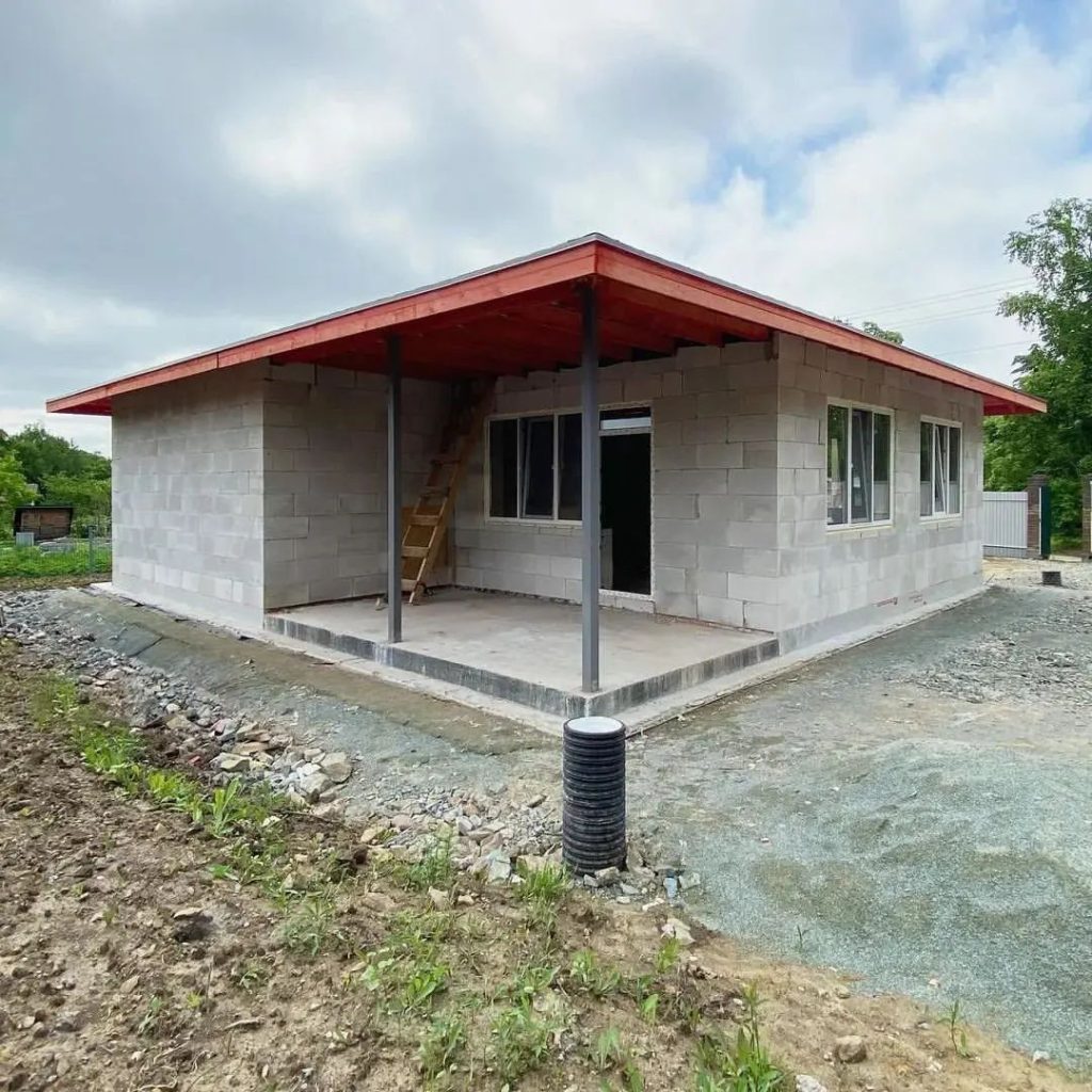 Photo of a small house made of concrete blocks with a wooden roof structure, surrounded by construction site.