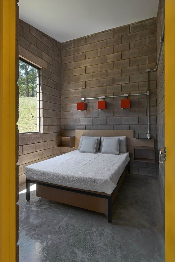 Interior of a minimalist bedroom featuring concrete block walls, a wooden bed frame, and natural light coming through the window.
